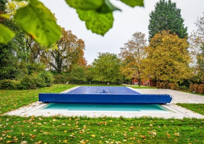 Chez-Elizabeth-et-Jacques-1 Une piscine couverte par une bâche bleue est entourée de pelouse verte et de grands arbres aux feuilles d'automne.