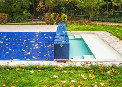 Chez-Elizabeth-et-Jacques-6 Une piscine partiellement couverte par une bâche bleue, entourée de pelouse verte et de feuilles d'automne dispersées.