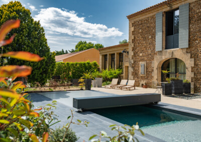 Een moderne tuin met zwembad, ligstoelen, groene planten en een stenen huis onder een blauwe lucht met wolken.