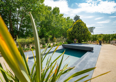 Een zwembad met een grijze afdekking, omgeven door groene bomen en planten, met een stenen terras ernaast.