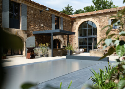 Een moderne patio met grijze overkapping naast een stenen gebouw met grote ramen en planten in potten op de grond.