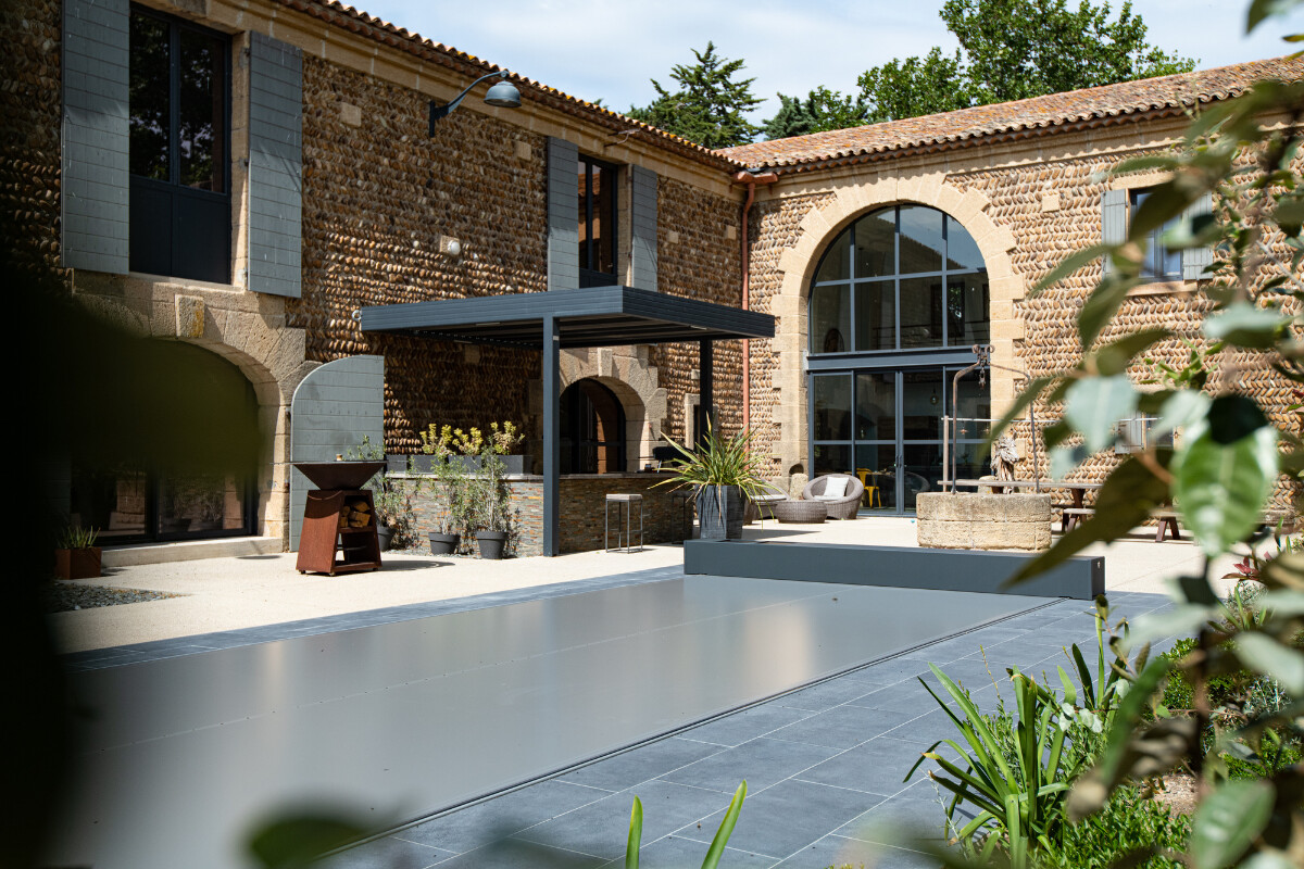 Een moderne patio met grijze overkapping naast een stenen gebouw met grote ramen en planten in potten op de grond.