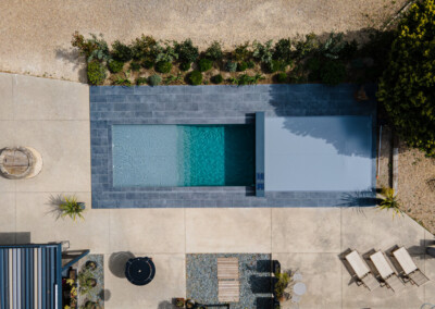 Zwembad met grijze tegels en afdekking, omringd door stenen terras, planten en tuinmeubilair, gefotografeerd vanuit de lucht.