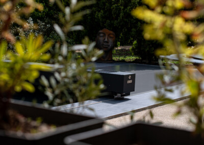 Een metalen zwembadafdekking in een tuin met groene planten en een boeddhabeeld op de achtergrond.