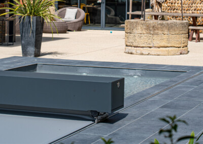 Grijze zwembadafdekking naast plantenbak en stenen tafel op een terras met stoelen op de achtergrond.